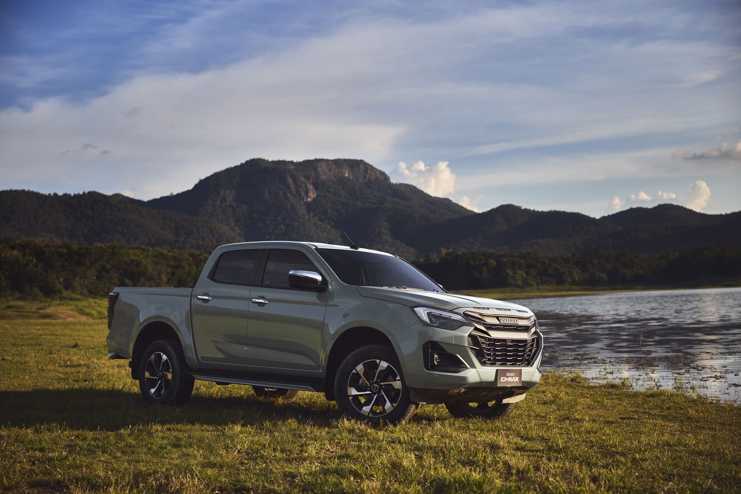 An ISUZU D-MAX parked on a grassy field by a lake, with mountains and a blue sky in the background.