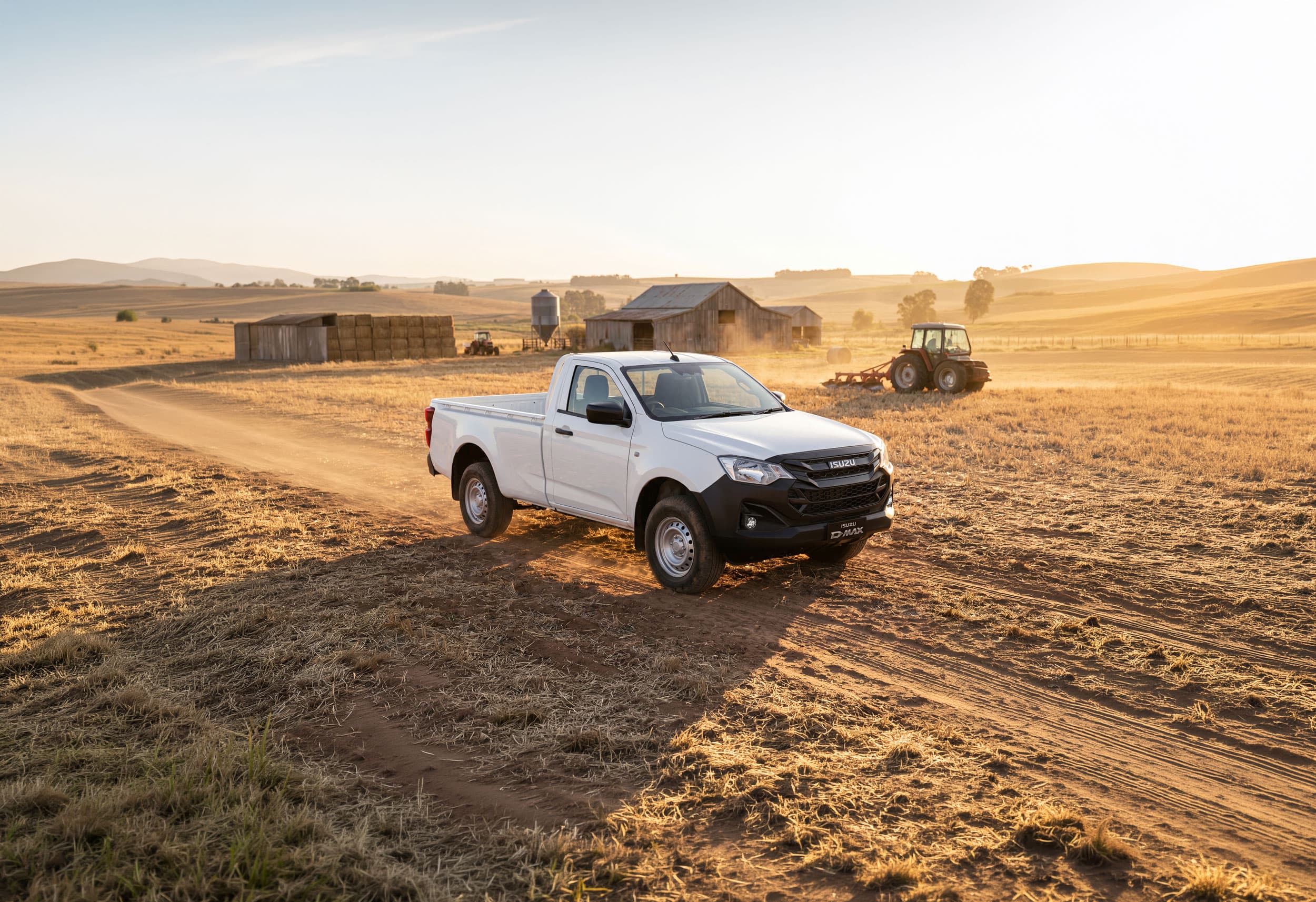 A white pickup truck drives on a dirt path through a sunlit farm field with hay bales, barns, and a tractor in the background.