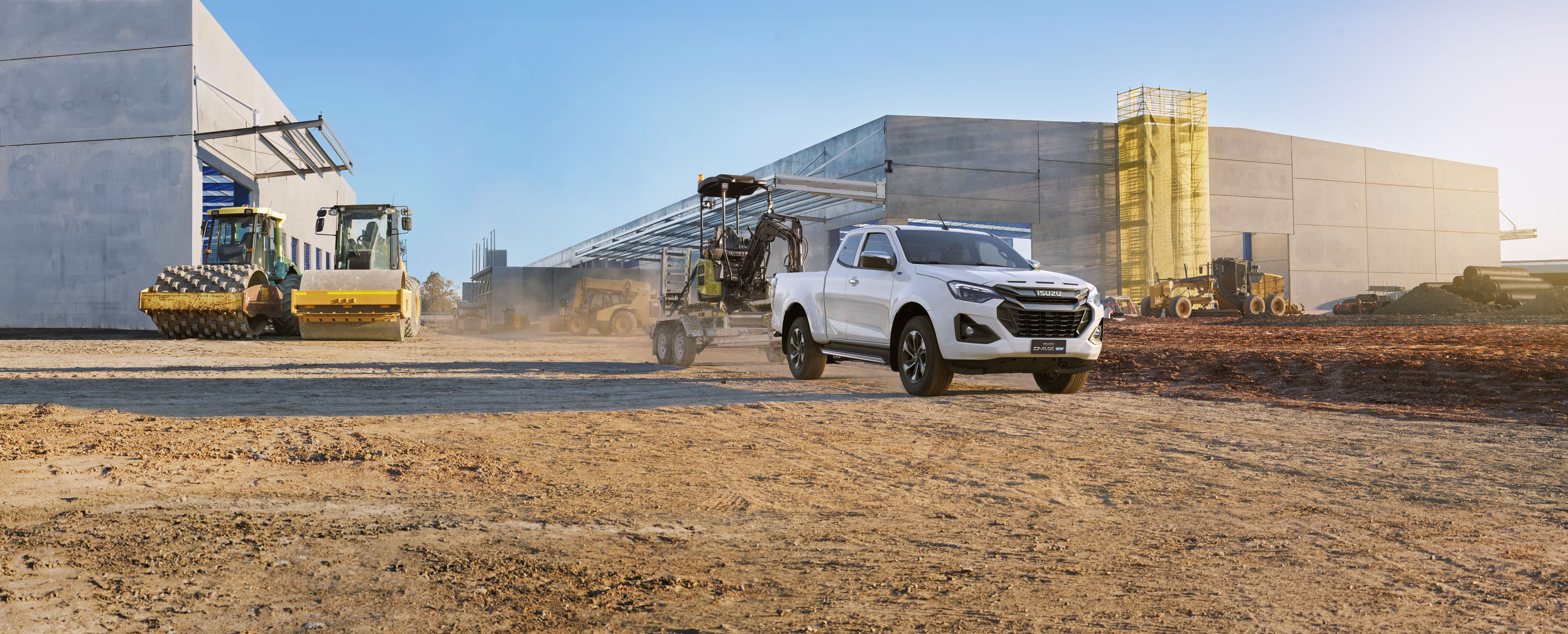 A white ISUZU D-MAX EV drives on a dusty construction site, with industrial buildings and machinery in the background under a clear blue sky.